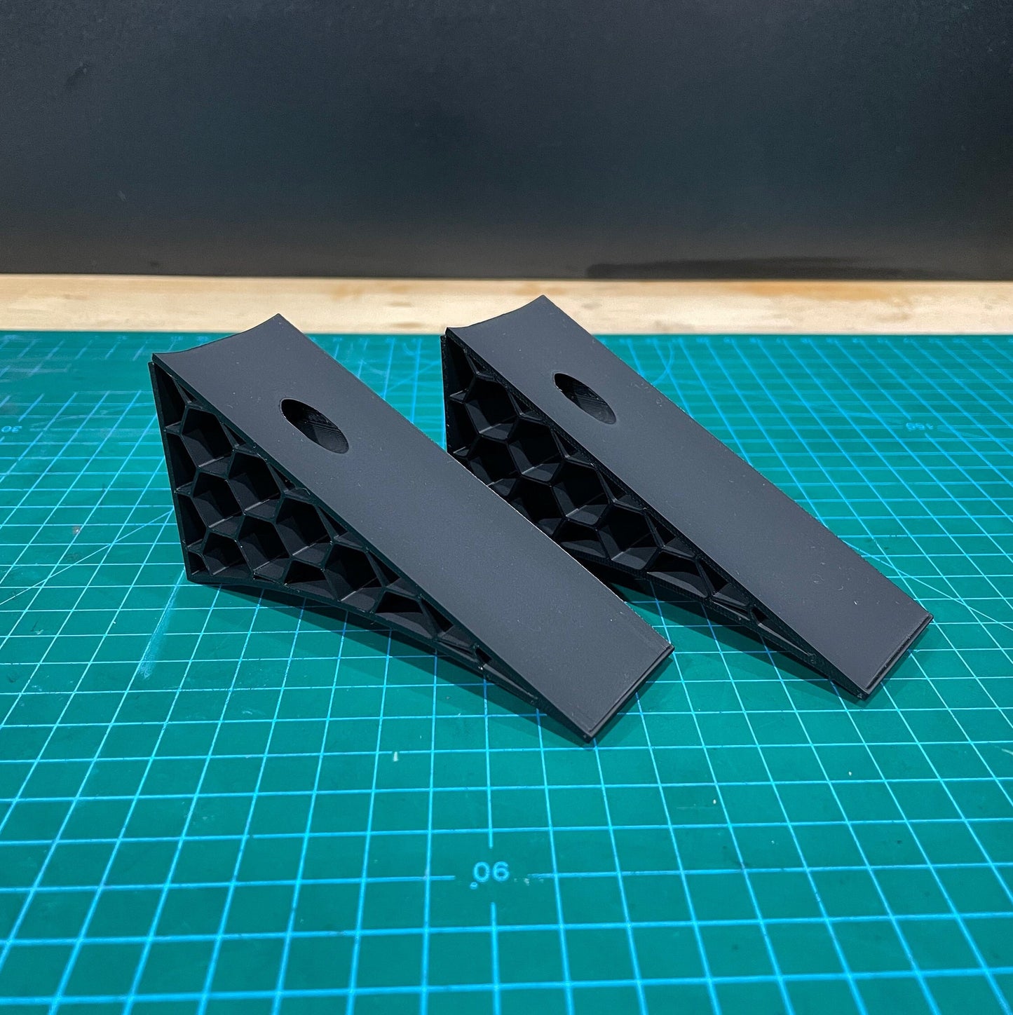 Pair of black 3D printed angled wall-mounted display brackets for 1:18 scale model cars, shown on a green cutting mat with honeycomb side structure visible.