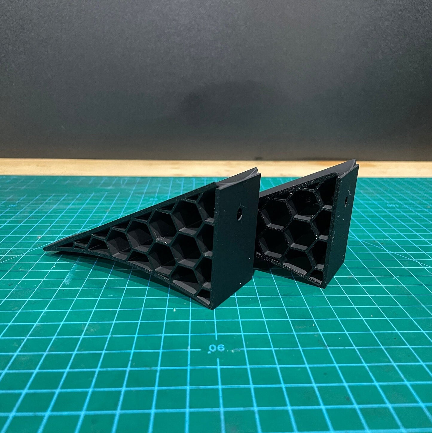 Pair of black 3D printed angled wall-mounted display brackets for 1:18 scale model cars, shown on a green cutting mat with honeycomb side structure visible.