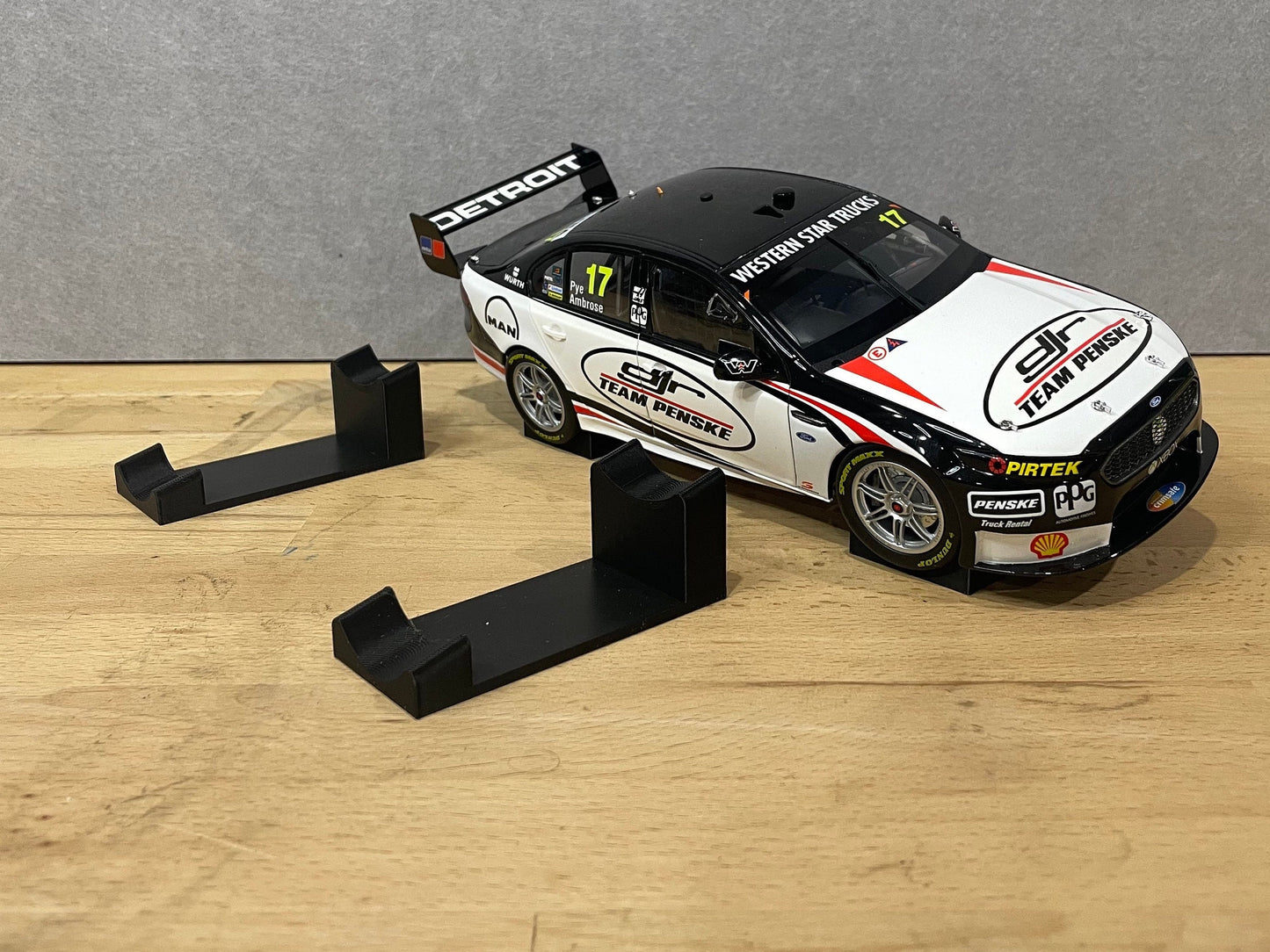 A 1:18 scale model of a V8 Supercar displayed on two black angled display mounts placed on a wooden surface.