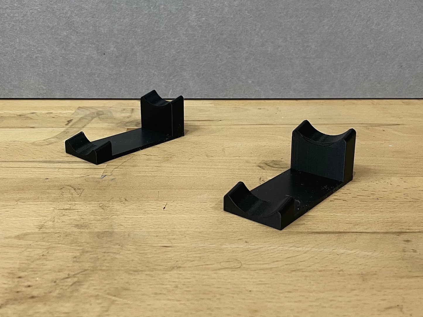 Pair of black 3D printed flat display mounts for 1:18 scale model cars, shown on a wooden surface, designed to cradle the front and rear axles securely.