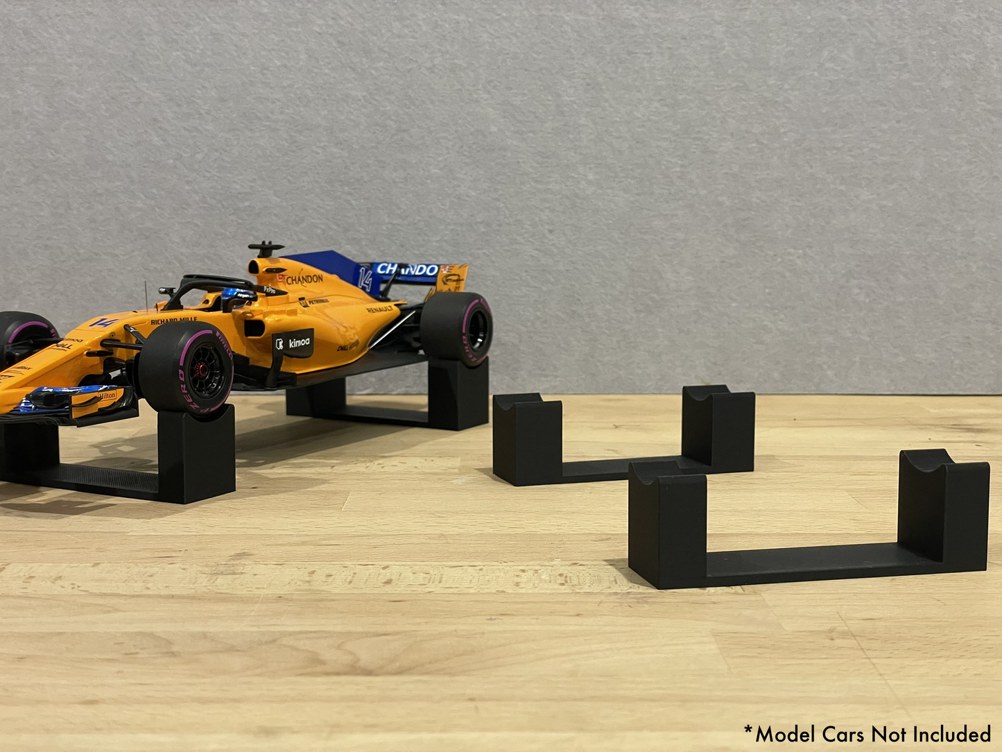 1:18 scale Formula 1 model car displayed on black 3D printed level display riser mounts, shown on a wooden surface against a grey background. The mounts are designed to cradle the front and rear axles securely for a clean, elevated presentation.