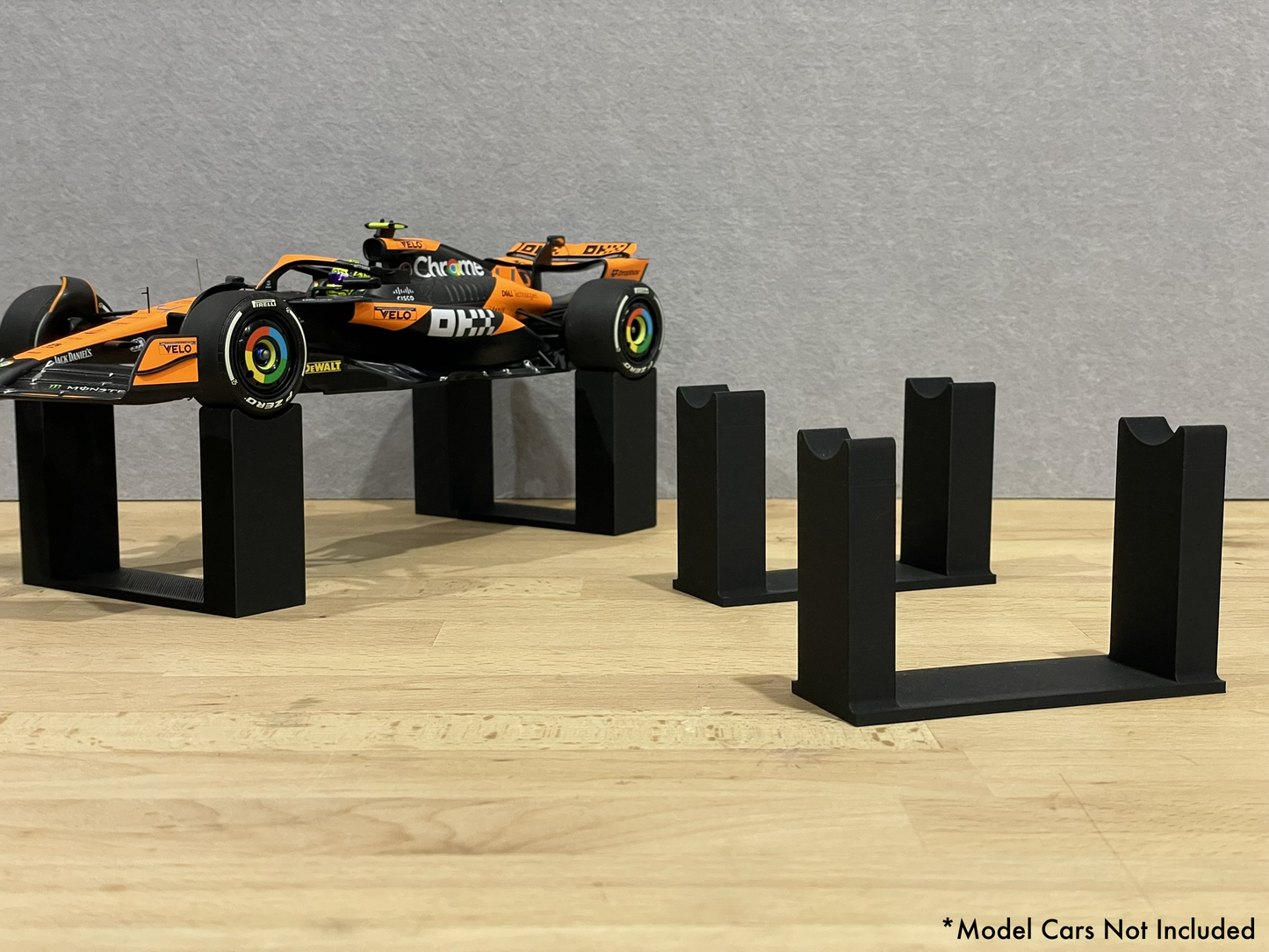 1:18 scale Formula 1 model car displayed on black 3D printed level display riser mounts, shown on a wooden surface against a grey background. The mounts are designed to cradle the front and rear axles securely for a clean, elevated presentation.