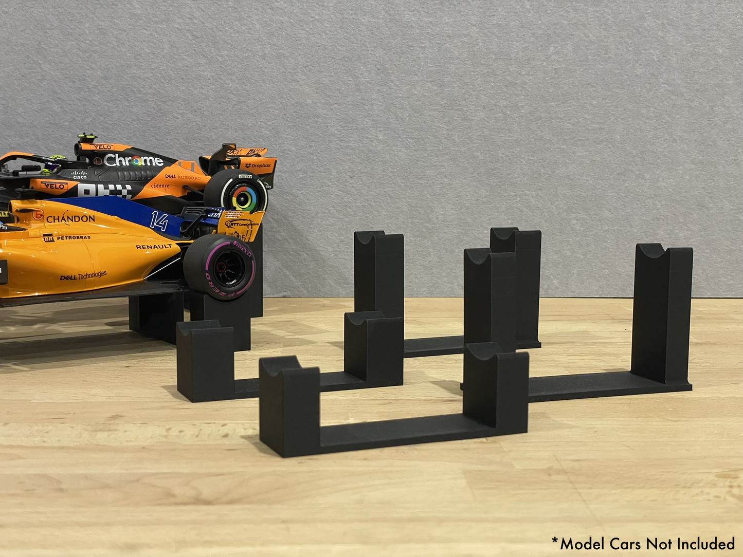1:18 scale Formula 1 model cars displayed on black 3D printed level display riser mounts, shown on a wooden surface against a grey background. The mounts are designed to cradle the front and rear axles securely for a clean, elevated presentation.