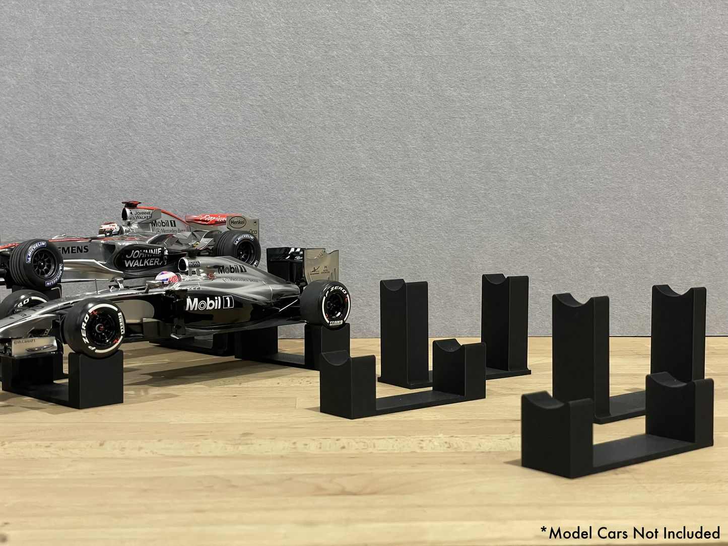 1:18 scale Formula 1 model cars displayed on black 3D printed level display riser mounts, shown on a wooden surface against a grey background. The mounts are designed to cradle the front and rear axles securely for a clean, elevated presentation.