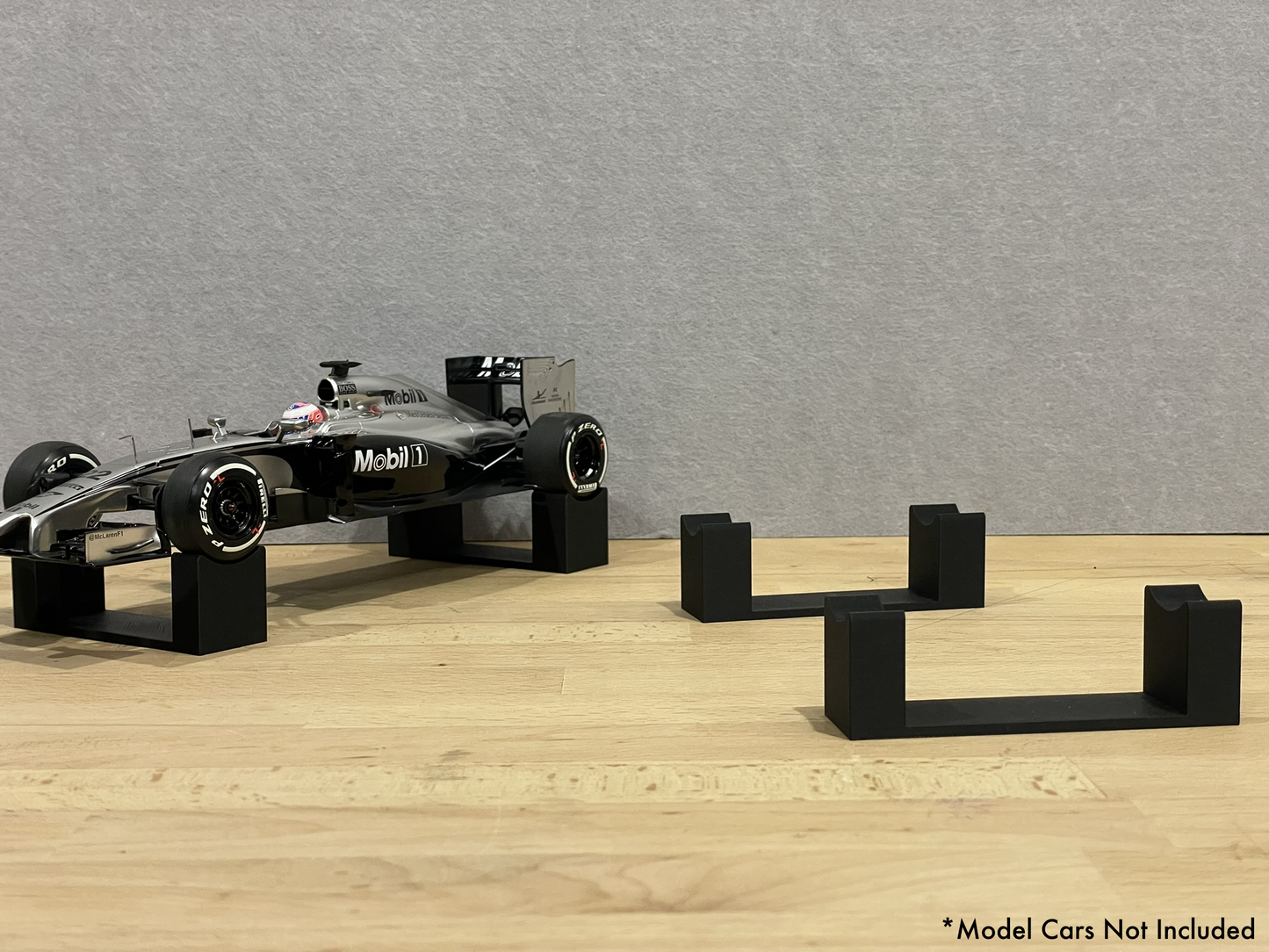 1:18 scale Formula 1 model car displayed on black 3D printed level display riser mounts, shown on a wooden surface against a grey background. The mounts are designed to cradle the front and rear axles securely for a clean, elevated presentation.
