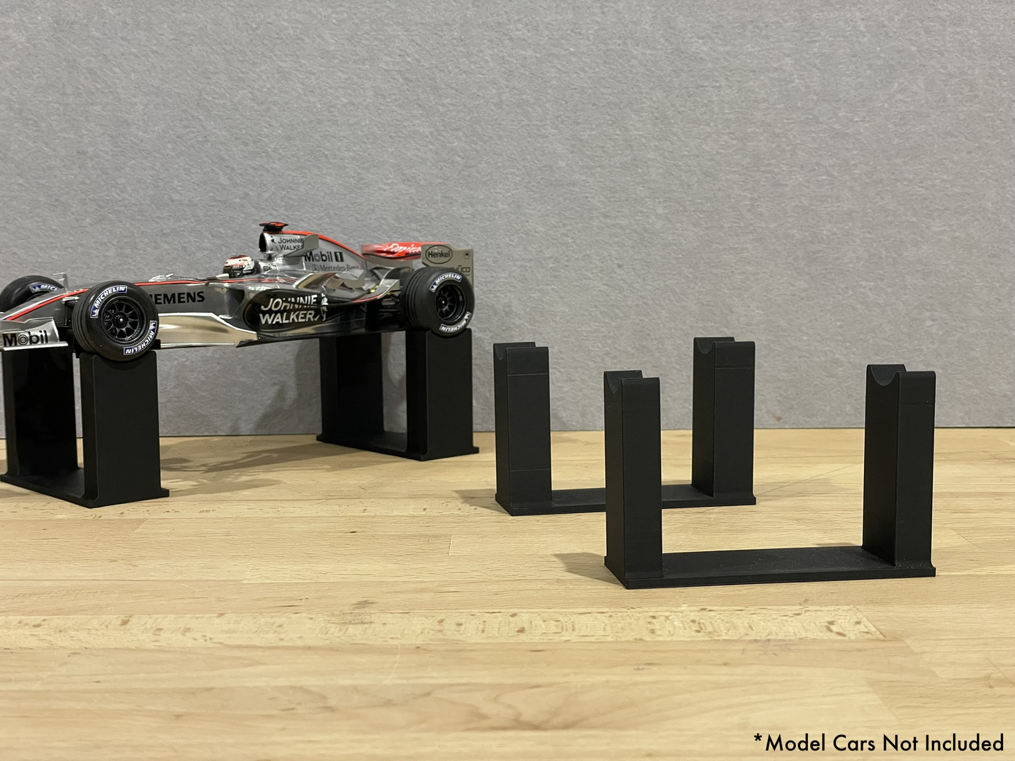 1:18 scale Formula 1 model car displayed on black 3D printed level display riser mounts, shown on a wooden surface against a grey background. The mounts are designed to cradle the front and rear axles securely for a clean, elevated presentation.