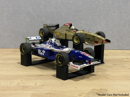 1:18 scale Formula 1 model cars displayed on black 3D printed level display riser mounts, shown on a wooden surface against a grey background. The mounts are designed to cradle the front and rear axles securely for a clean, elevated presentation.