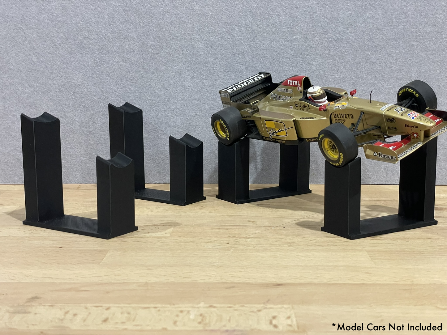 1:18 scale Formula 1 model car displayed on black 3D printed angled display mounts, shown on a wooden surface against a grey background. The mounts are designed to cradle the front and rear axles securely for a clean, elevated presentation.