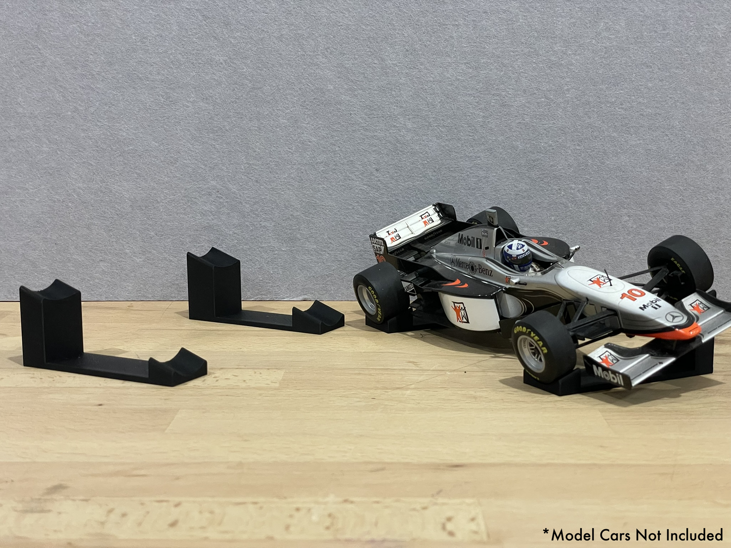 1:18 scale Formula 1 model car displayed on black 3D printed angled display mounts, shown on a wooden surface against a grey background. The mounts are designed to cradle the front and rear axles securely for a clean, elevated presentation.