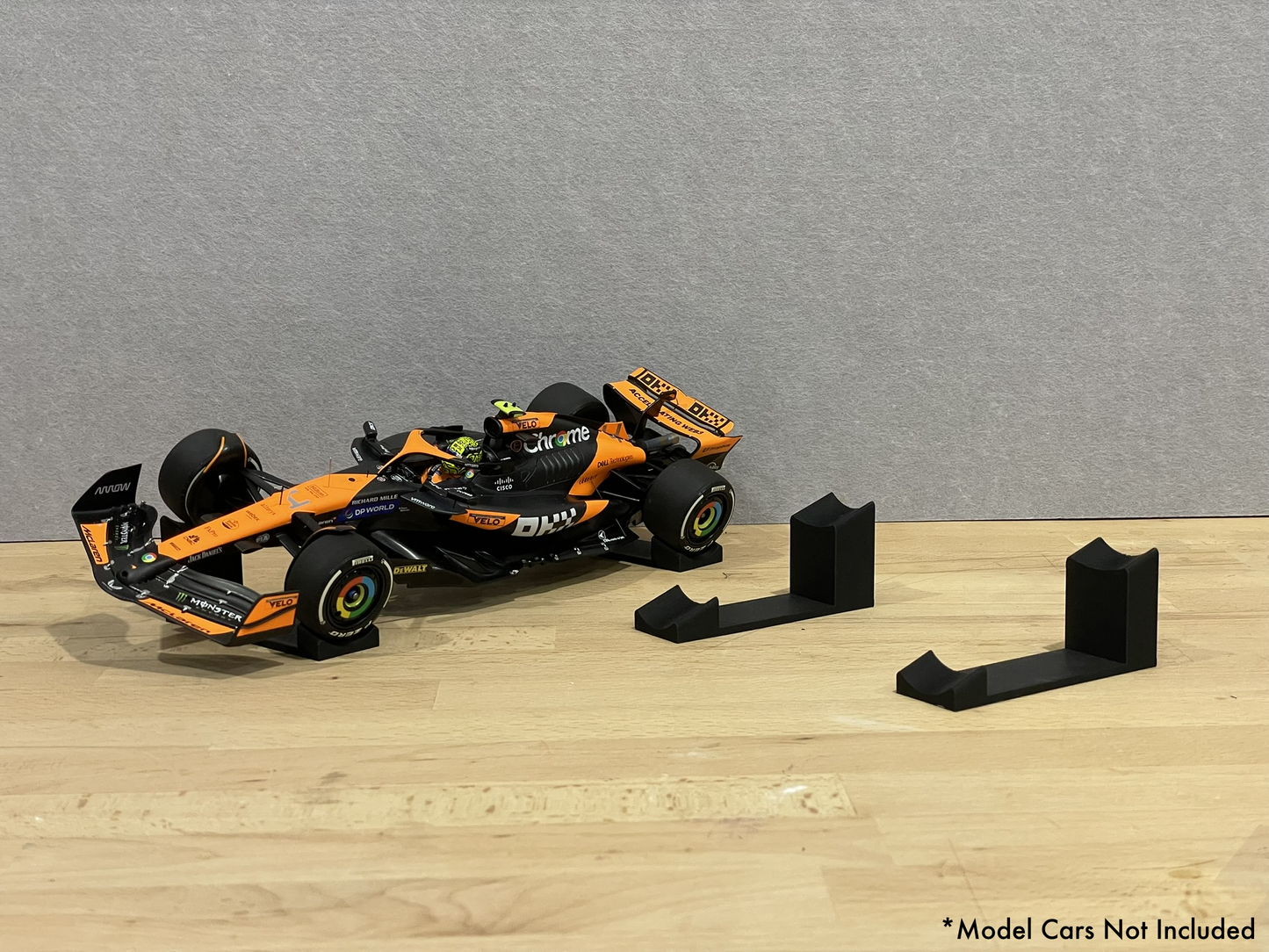 1:18 scale Formula 1 model car displayed on black 3D printed angled display mounts, shown on a wooden surface against a grey background. The mounts are designed to cradle the front and rear axles securely for a clean, elevated presentation.