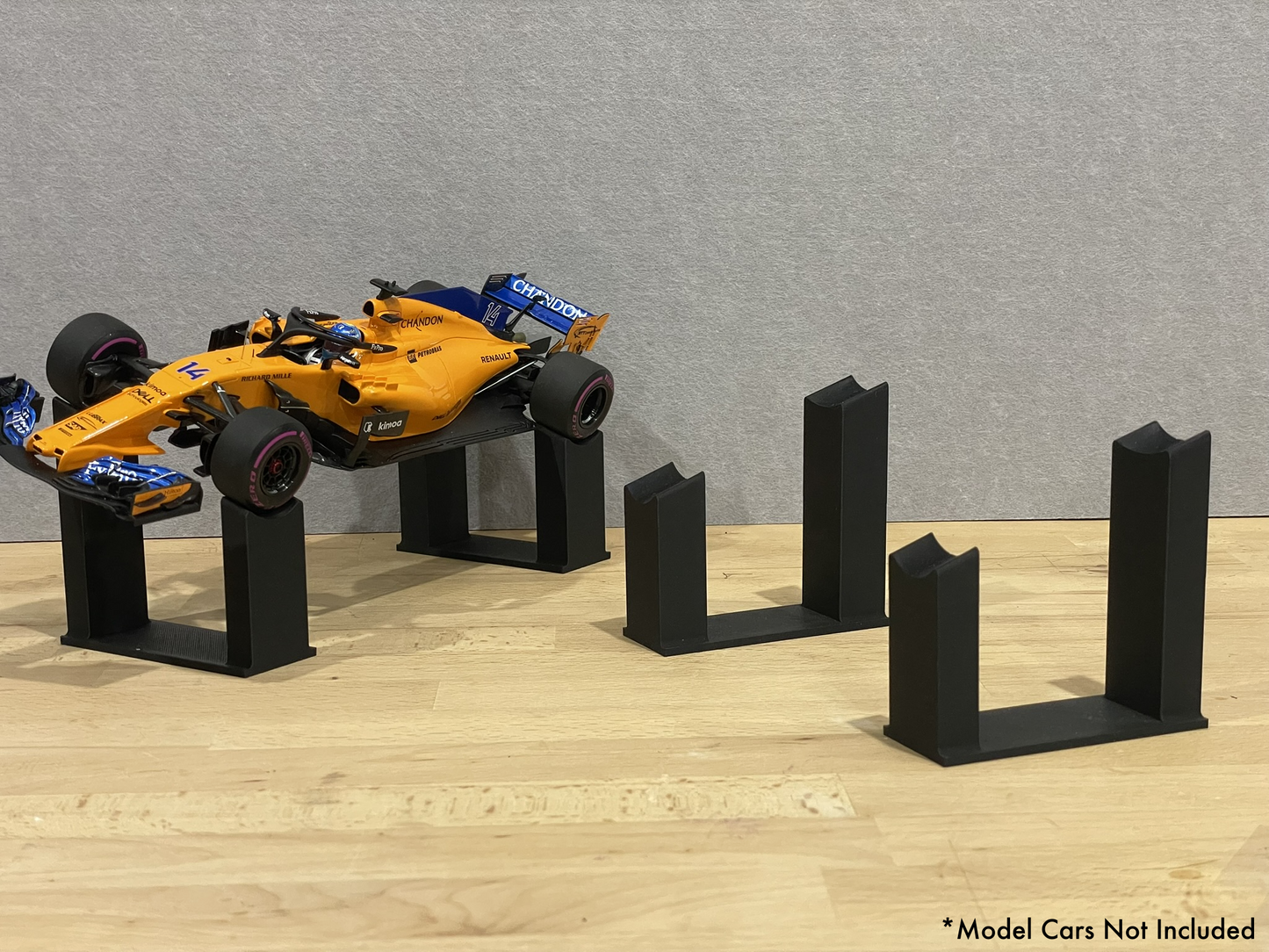1:18 scale Formula 1 model car displayed on black 3D printed angled display mounts, shown on a wooden surface against a grey background. The mounts are designed to cradle the front and rear axles securely for a clean, elevated presentation.