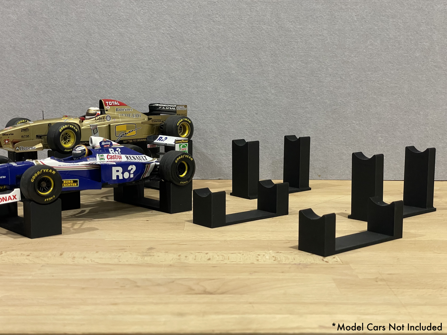 Set of black 3D printed level display mounts for 1:18 scale model formula 1 cars, shown on a wooden surface, designed to cradle the front and rear axles securely.