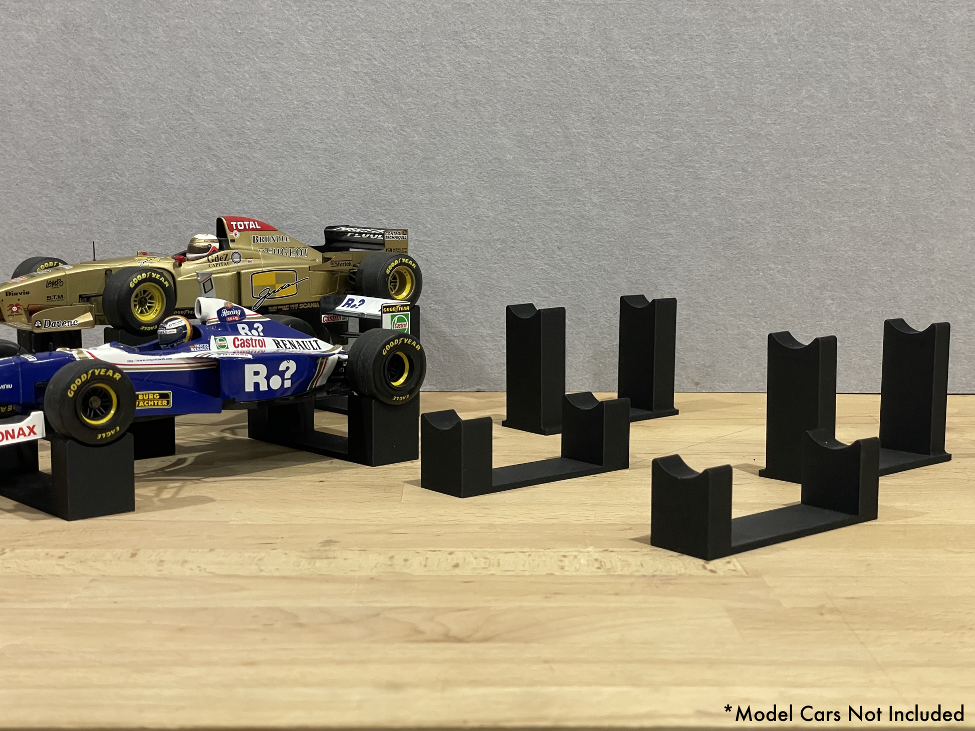 Set of black 3D printed level display mounts for 1:18 scale model formula 1 cars, shown on a wooden surface, designed to cradle the front and rear axles securely.