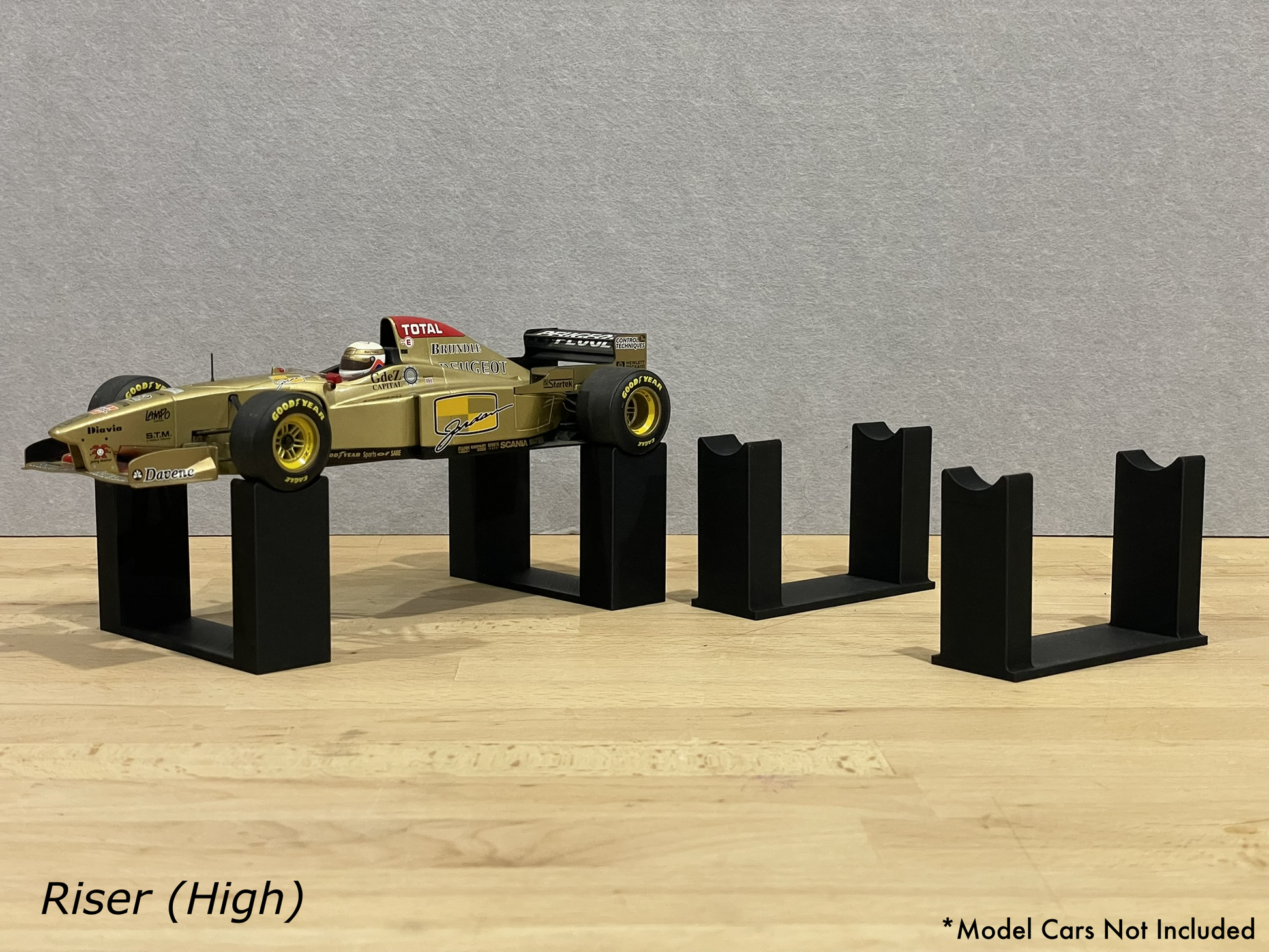 1:18 scale Formula 1 model car displayed on black 3D printed level display riser mounts, shown on a wooden surface against a grey background. The mounts are designed to cradle the front and rear axles securely for a clean, elevated presentation.