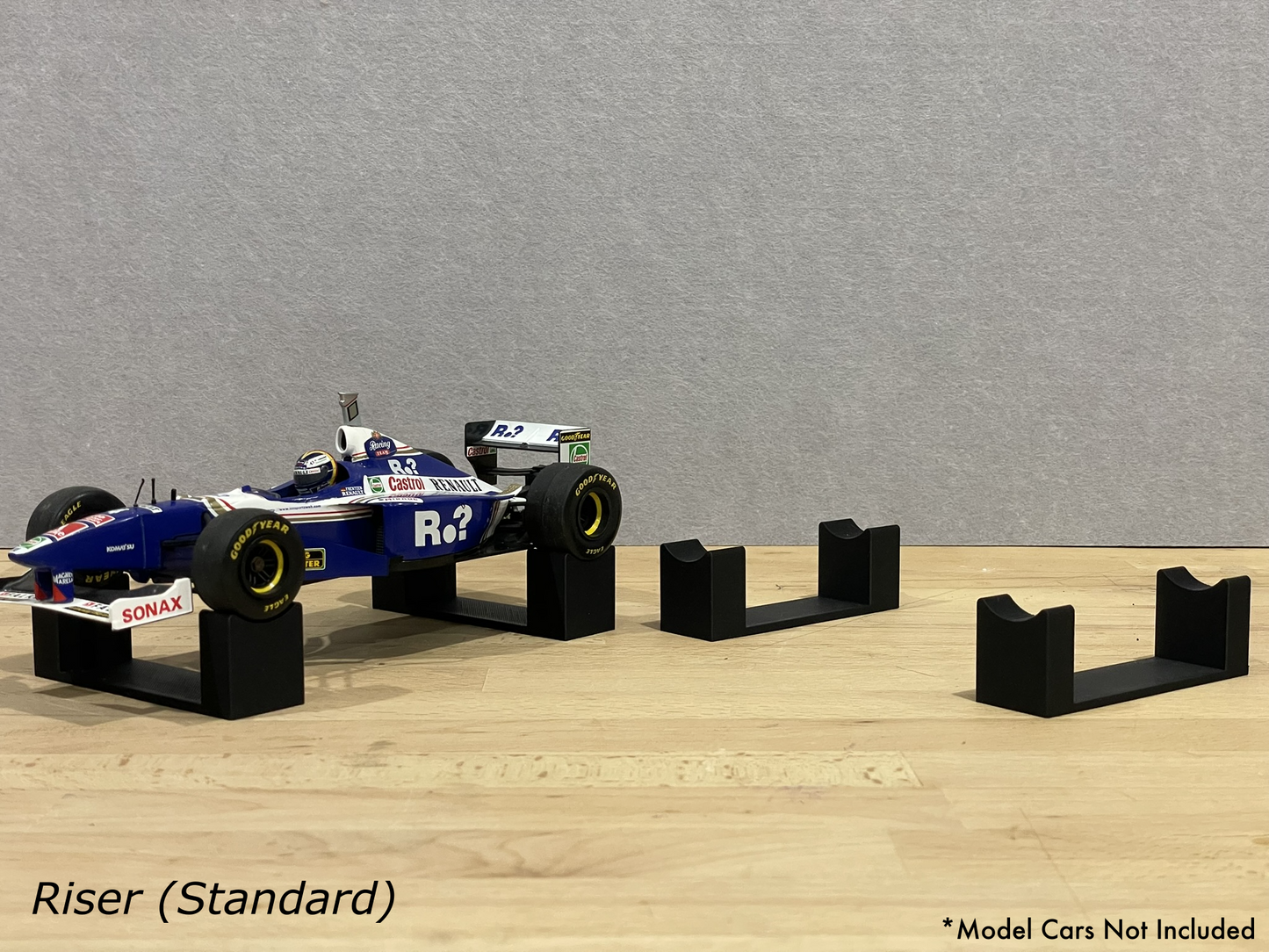 1:18 scale Formula 1 model car displayed on black 3D printed level display riser mounts, shown on a wooden surface against a grey background. The mounts are designed to cradle the front and rear axles securely for a clean, elevated presentation.