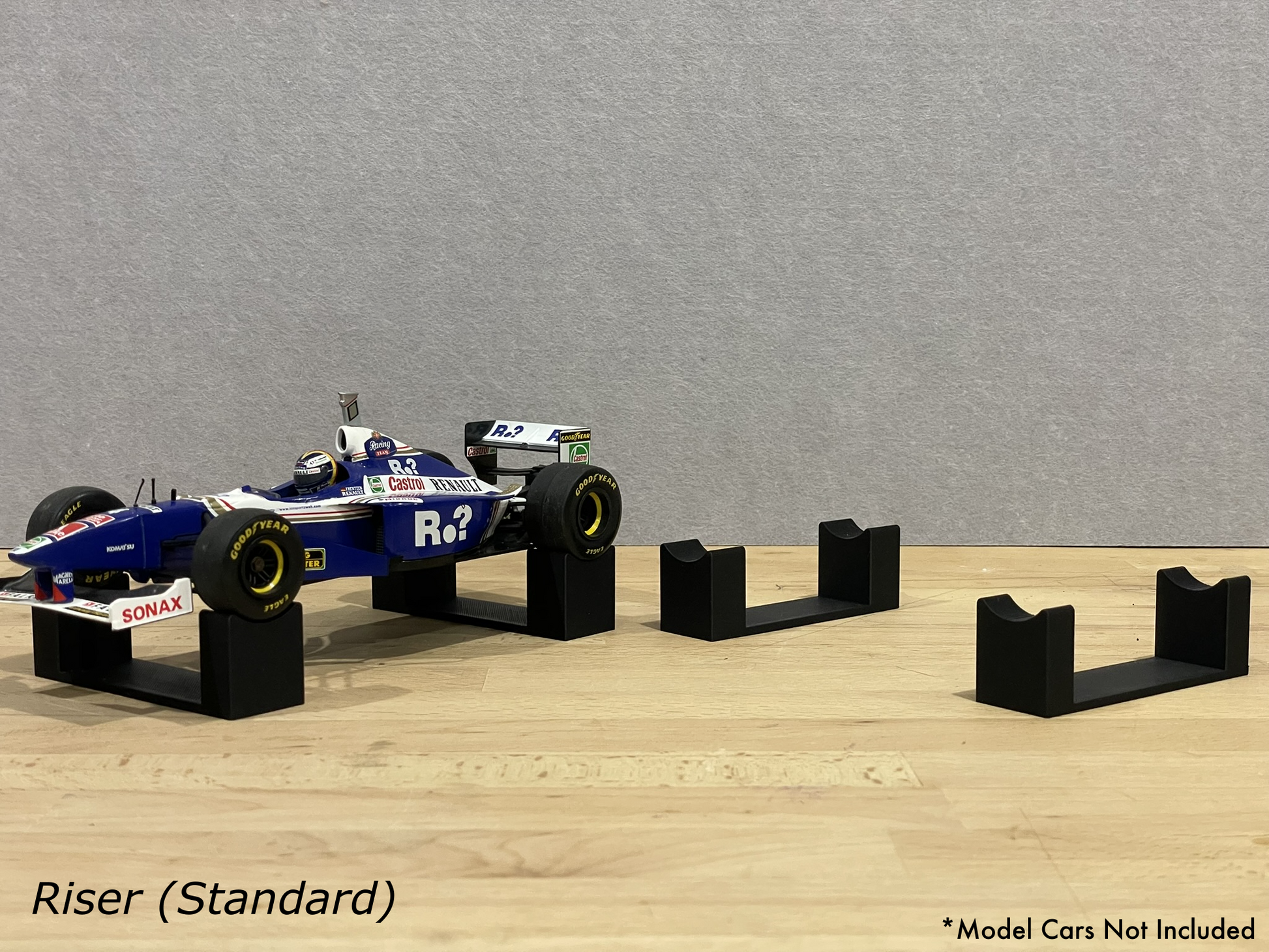 1:18 scale Formula 1 model car displayed on black 3D printed level display riser mounts, shown on a wooden surface against a grey background. The mounts are designed to cradle the front and rear axles securely for a clean, elevated presentation.