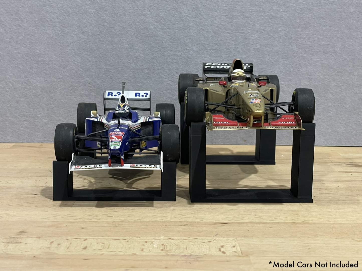 1:18 scale Formula 1 model cars displayed on black 3D printed level display riser mounts, shown on a wooden surface against a grey background. The mounts are designed to cradle the front and rear axles securely for a clean, elevated presentation.
