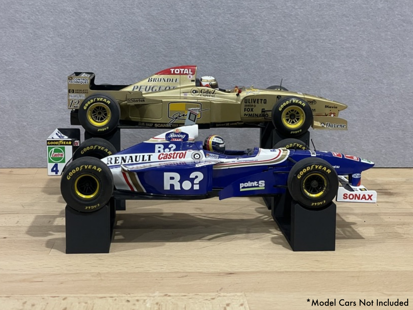 1:18 scale Formula 1 model cars displayed on black 3D printed level display riser mounts, shown on a wooden surface against a grey background. The mounts are designed to cradle the front and rear axles securely for a clean, elevated presentation.