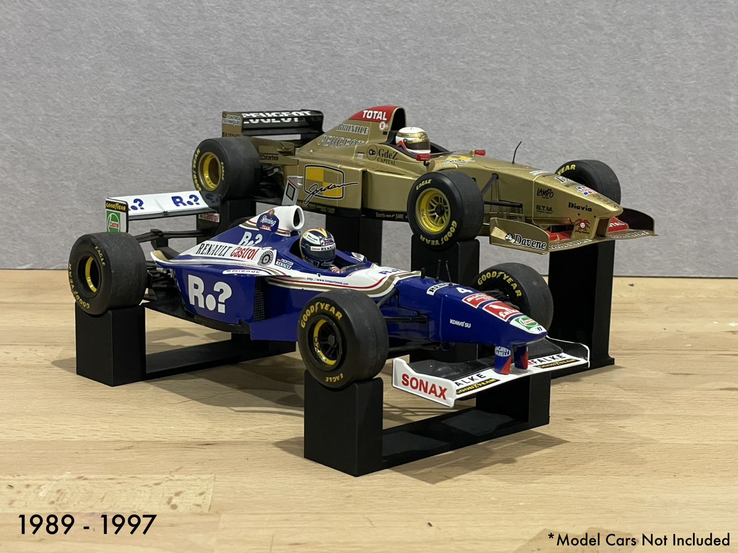 1:18 scale Formula 1 model cars displayed on black 3D printed level display riser mounts, shown on a wooden surface against a grey background. The mounts are designed to cradle the front and rear axles securely for a clean, elevated presentation.