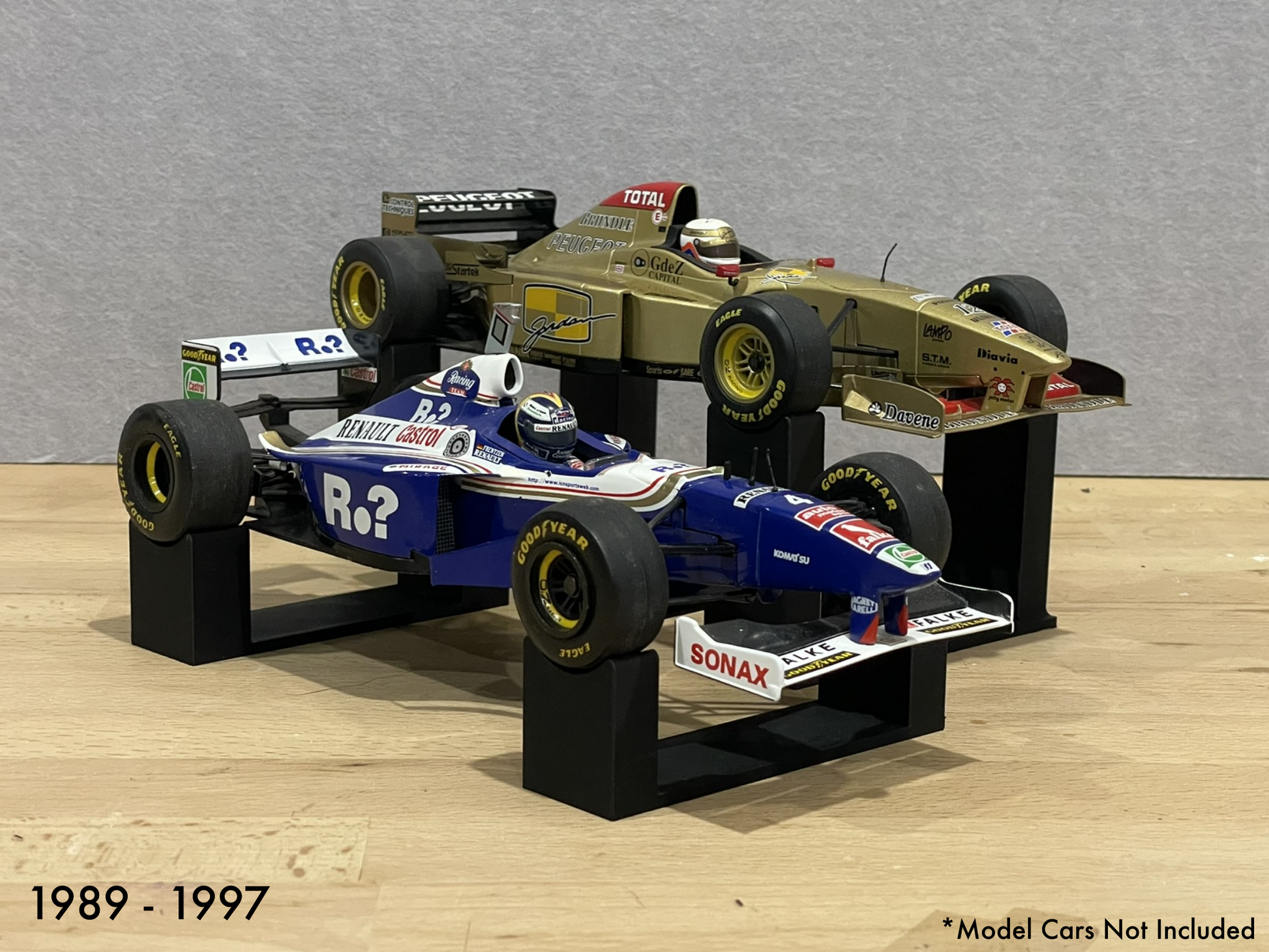1:18 scale Formula 1 model cars displayed on black 3D printed level display riser mounts, shown on a wooden surface against a grey background. The mounts are designed to cradle the front and rear axles securely for a clean, elevated presentation.