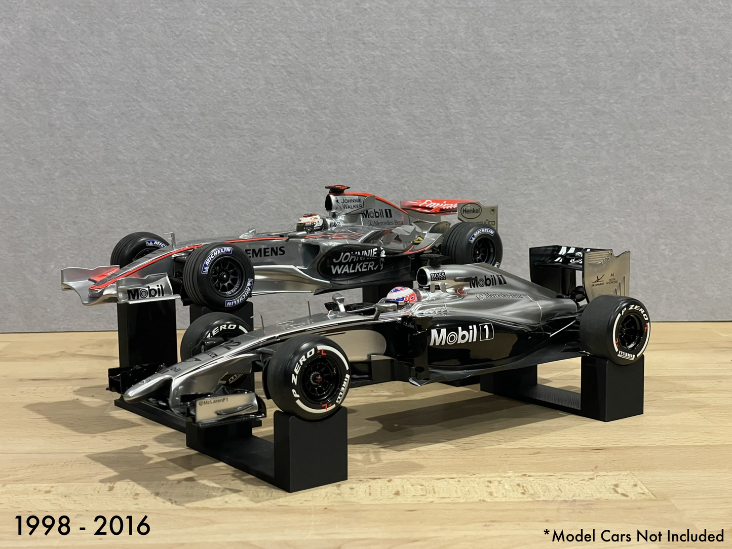 1:18 scale Formula 1 model cars displayed on black 3D printed level display riser mounts, shown on a wooden surface against a grey background. The mounts are designed to cradle the front and rear axles securely for a clean, elevated presentation.