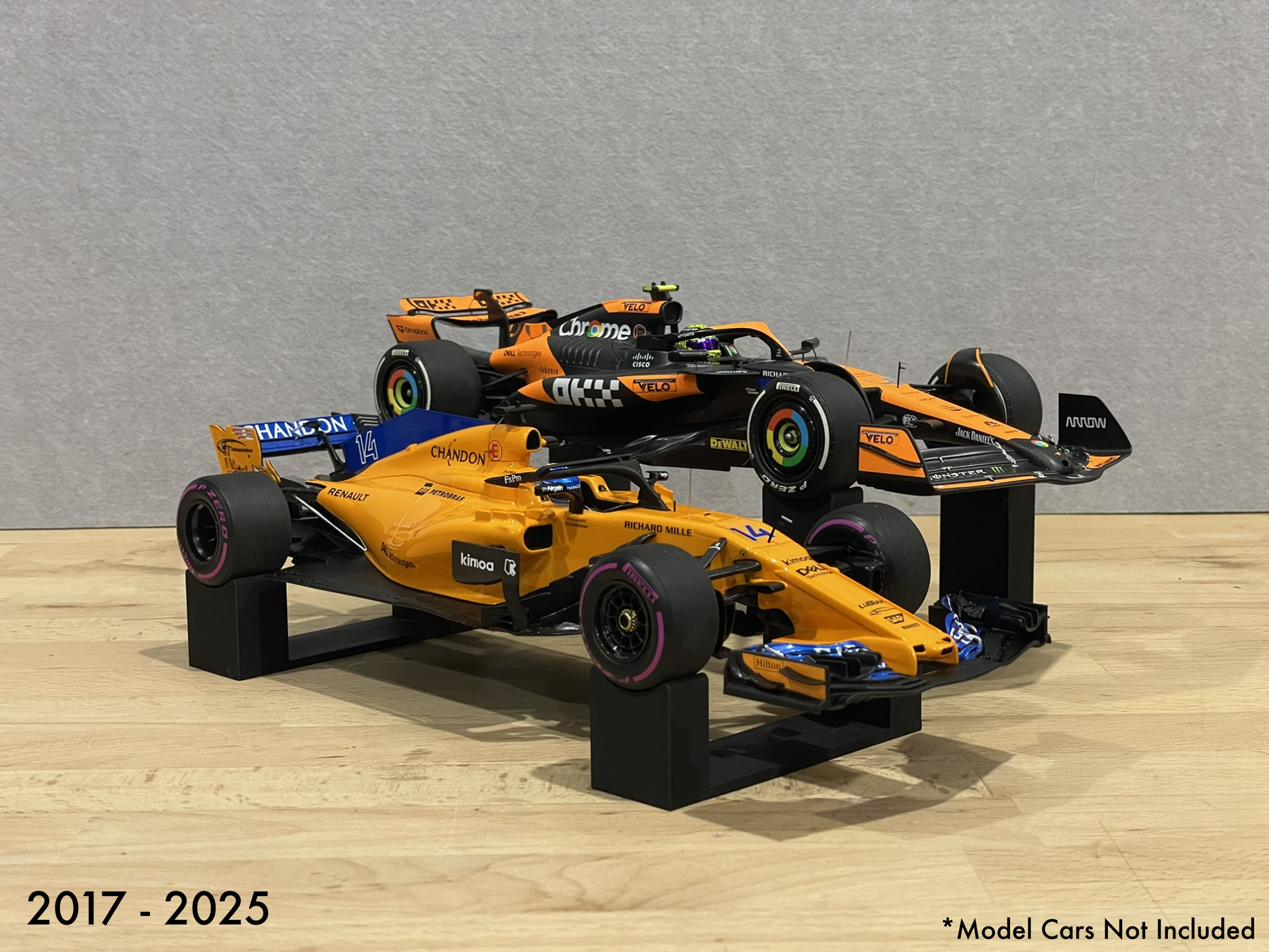 1:18 scale Formula 1 model cars displayed on black 3D printed level display riser mounts, shown on a wooden surface against a grey background. The mounts are designed to cradle the front and rear axles securely for a clean, elevated presentation.