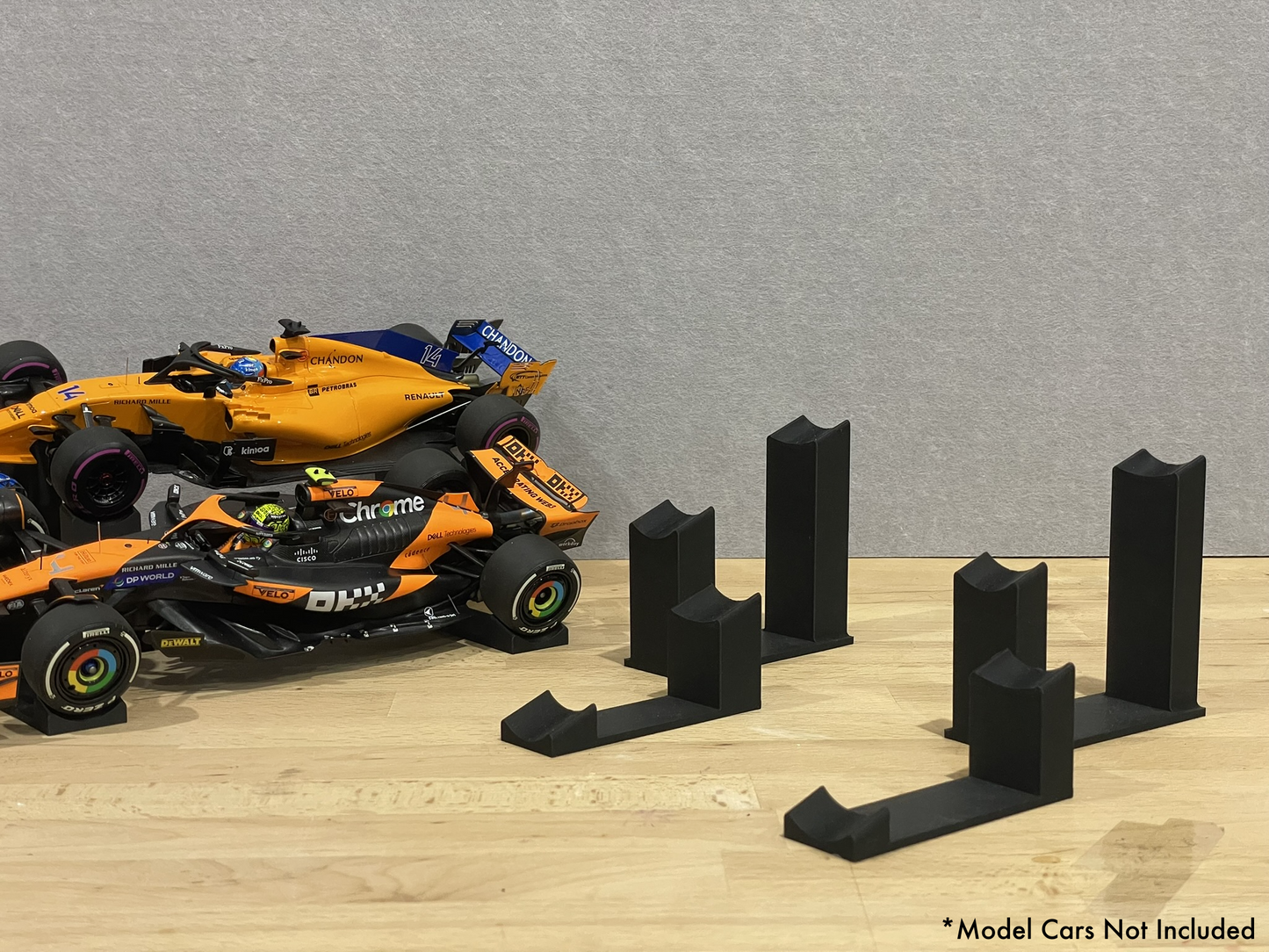 two 1:18 scale Formula 1 model cars displayed on black 3D printed angled display mounts, shown on a wooden surface against a grey background. The mounts are designed to cradle the front and rear axles securely for a clean, elevated presentation.