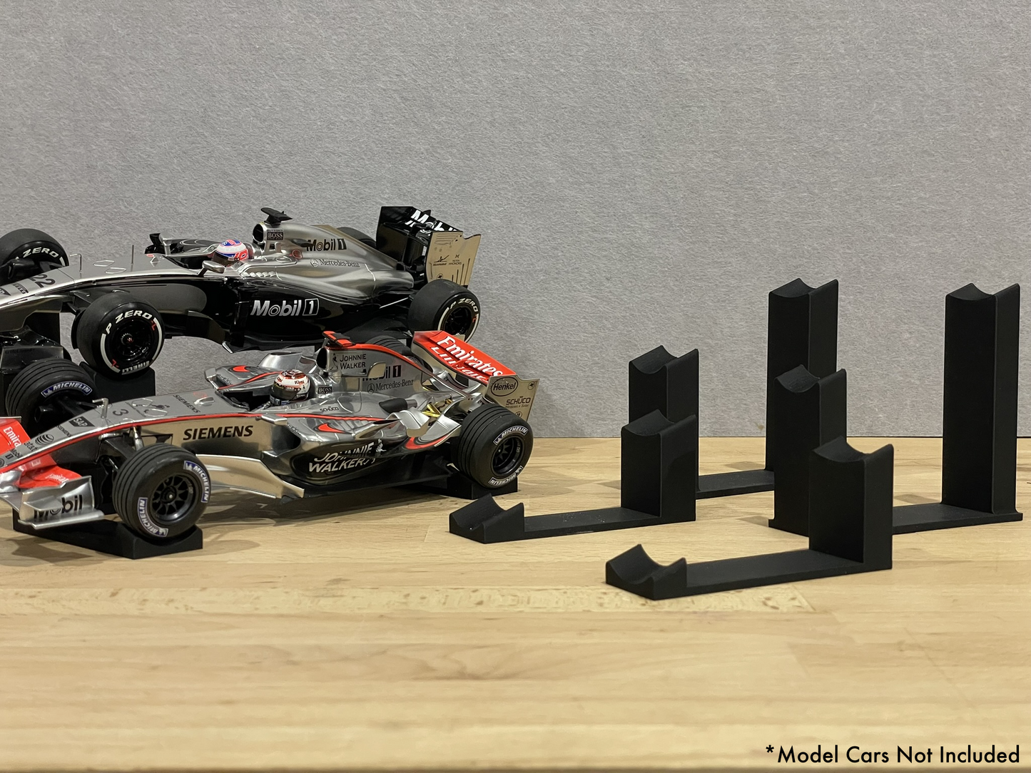 Two 1:18 scale Formula 1 model cars displayed on black 3D printed angled display mounts, shown on a wooden surface against a grey background. The mounts are designed to cradle the front and rear axles securely for a clean, elevated presentation.