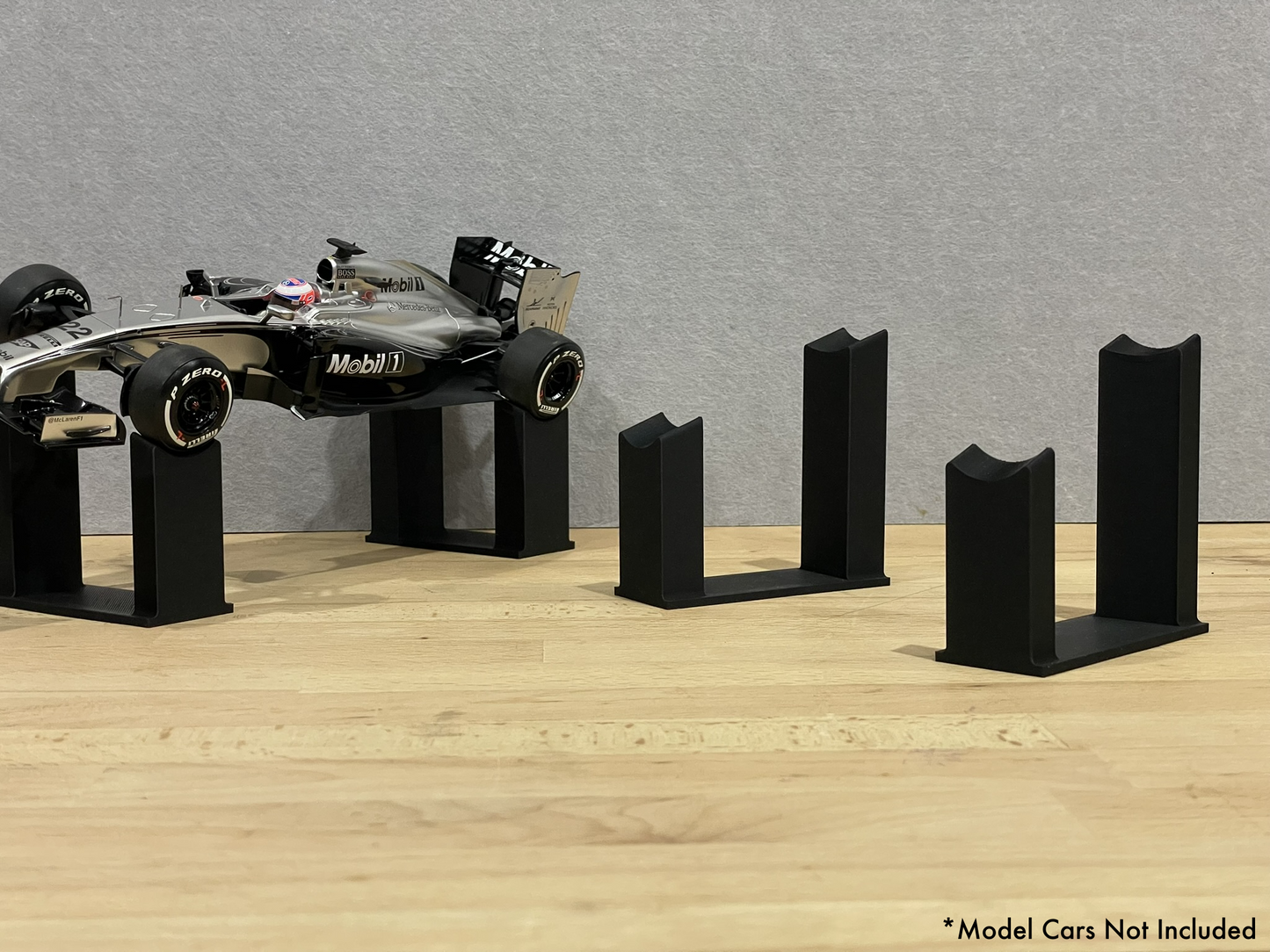 1:18 scale Formula 1 model car displayed on black 3D printed angled display mounts, shown on a wooden surface against a grey background. The mounts are designed to cradle the front and rear axles securely for a clean, elevated presentation.