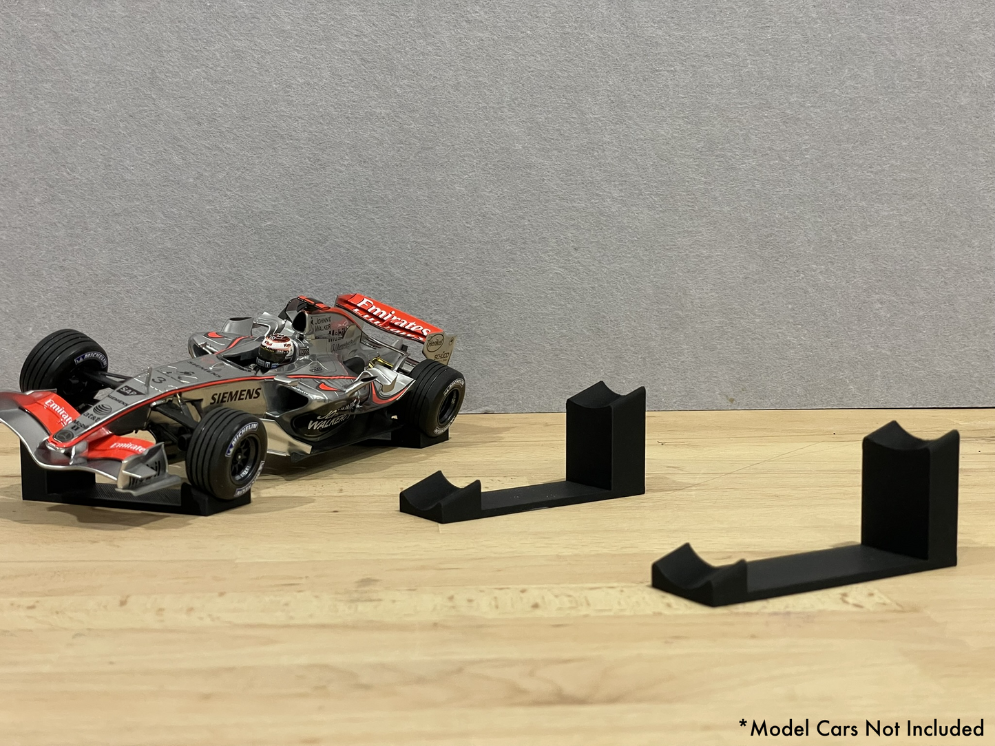 1:18 scale Formula 1 model car displayed on black 3D printed angled display mounts, shown on a wooden surface against a grey background. The mounts are designed to cradle the front and rear axles securely for a clean, elevated presentation.