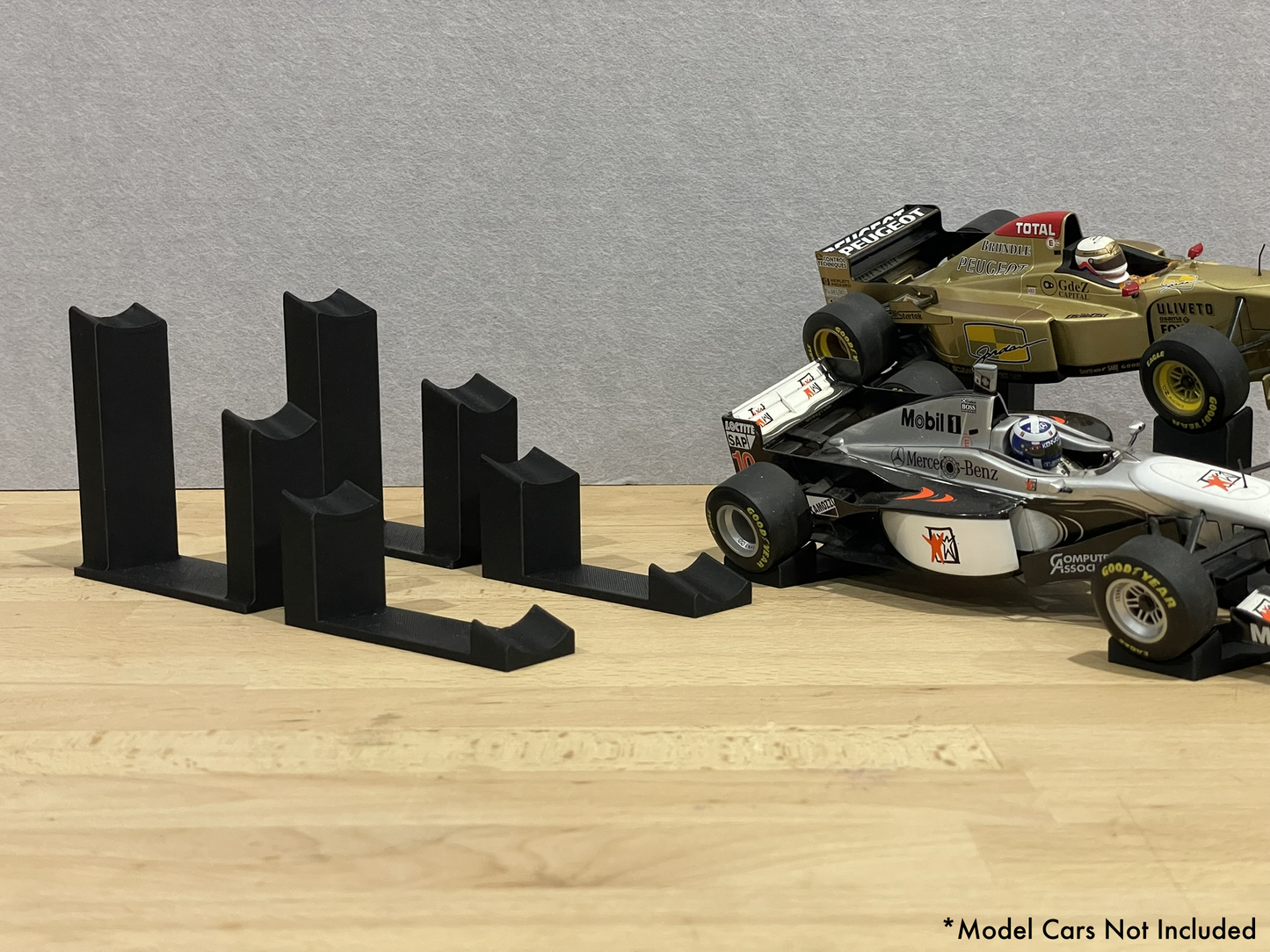 two 1:18 scale Formula 1 model cars displayed on black 3D printed angled display mounts, shown on a wooden surface against a grey background. The mounts are designed to cradle the front and rear axles securely for a clean, elevated presentation.