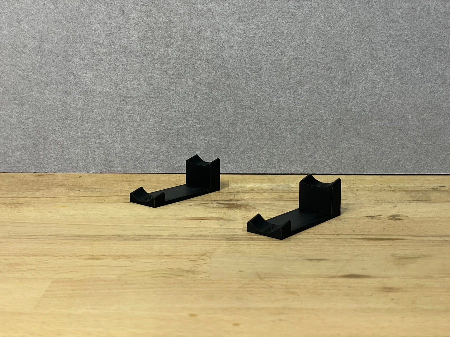 Pair of black 3D printed flat display mounts for 1:24 scale model cars, shown on a wooden surface, designed to cradle the front and rear axles securely.
