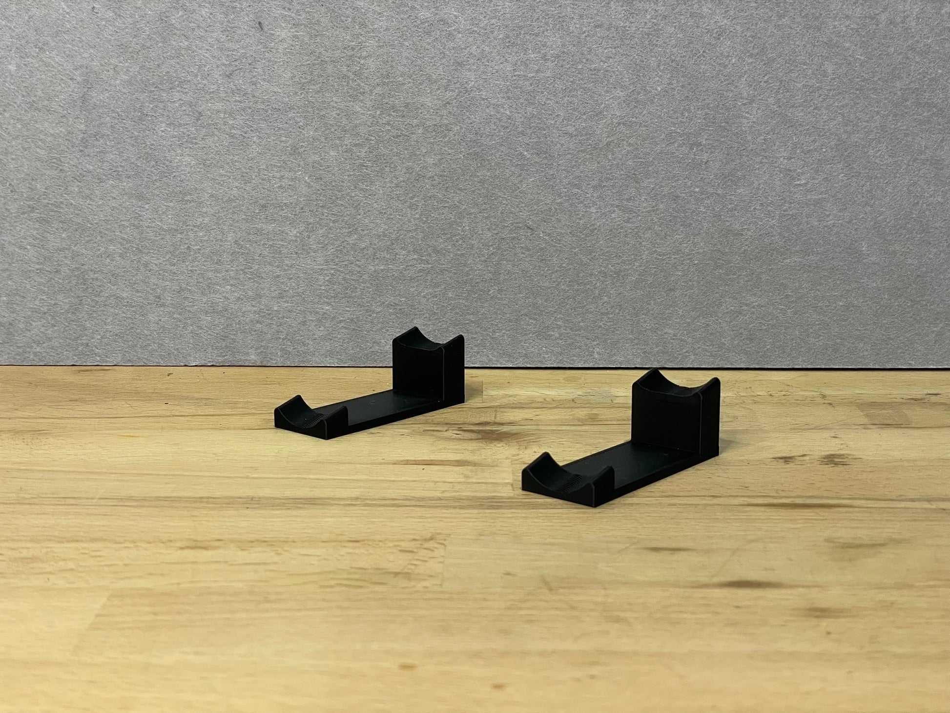 Pair of black 3D printed flat display mounts for 1:24 scale model cars, shown on a wooden surface, designed to cradle the front and rear axles securely.