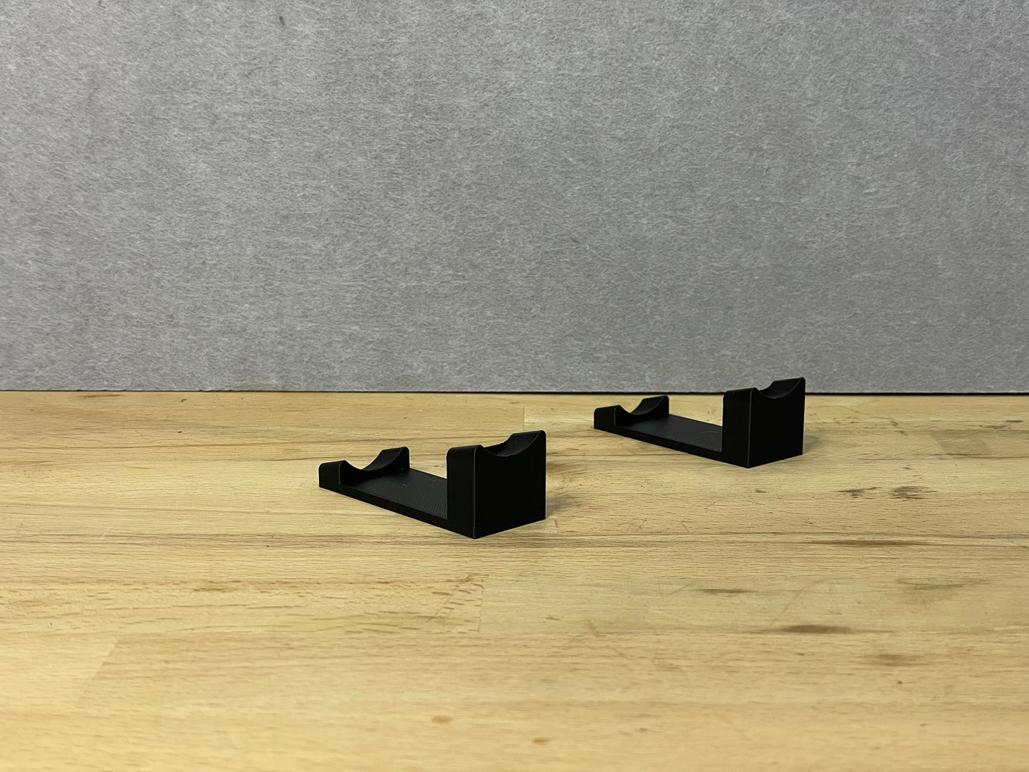 Pair of black 3D printed flat display mounts for 1:24 scale model cars, shown on a wooden surface, designed to cradle the front and rear axles securely.