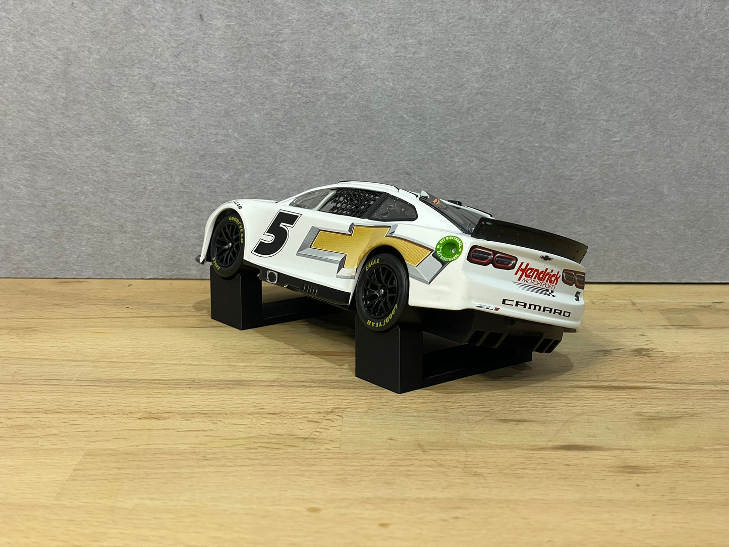 A 1:24 scale model of a NASCAR car displayed on two black angled display mounts on a wooden surface against a grey background.
