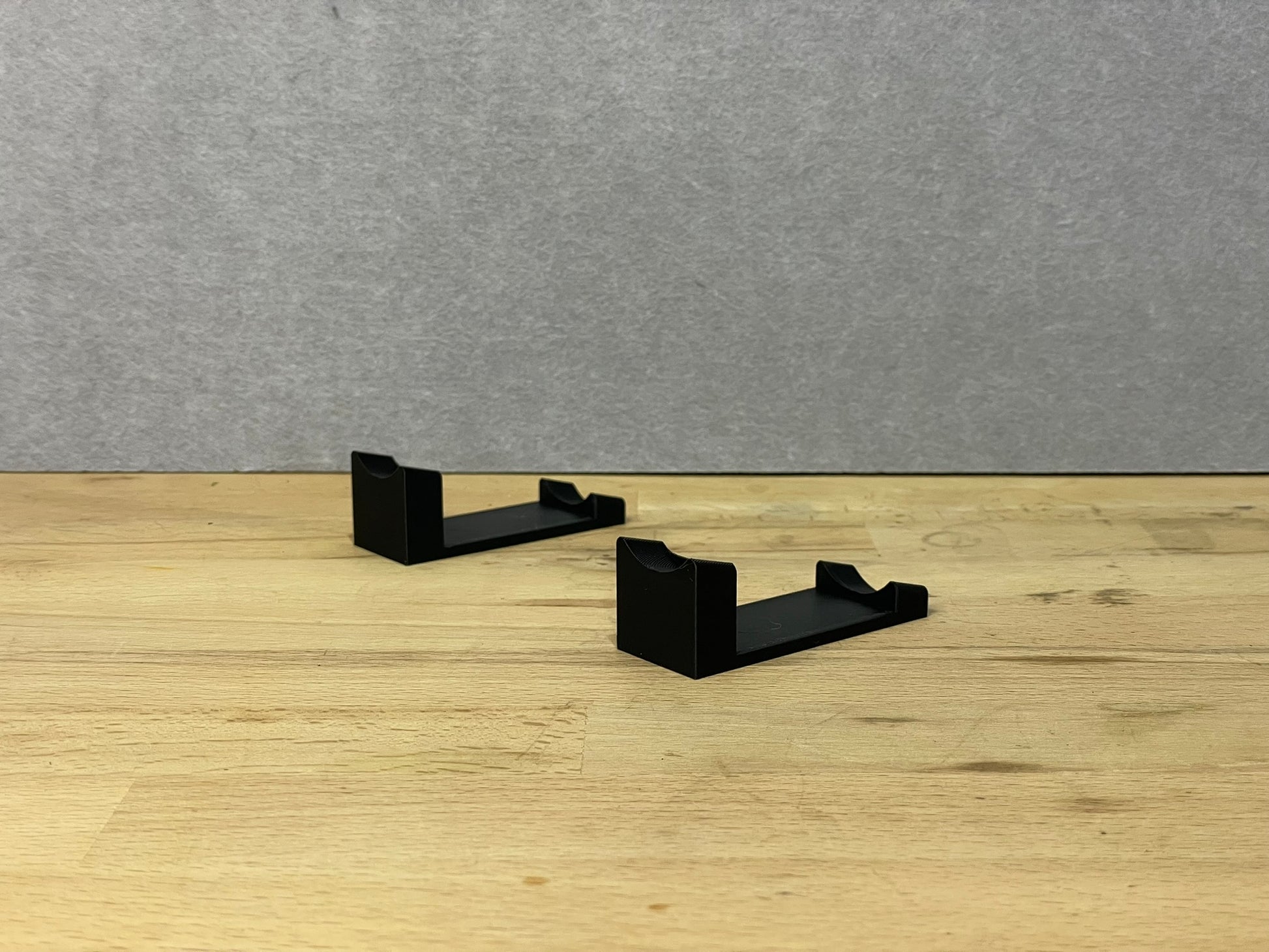 Pair of black 3D printed flat display mounts for 1:24 scale model cars, shown on a wooden surface, designed to cradle the front and rear axles securely.