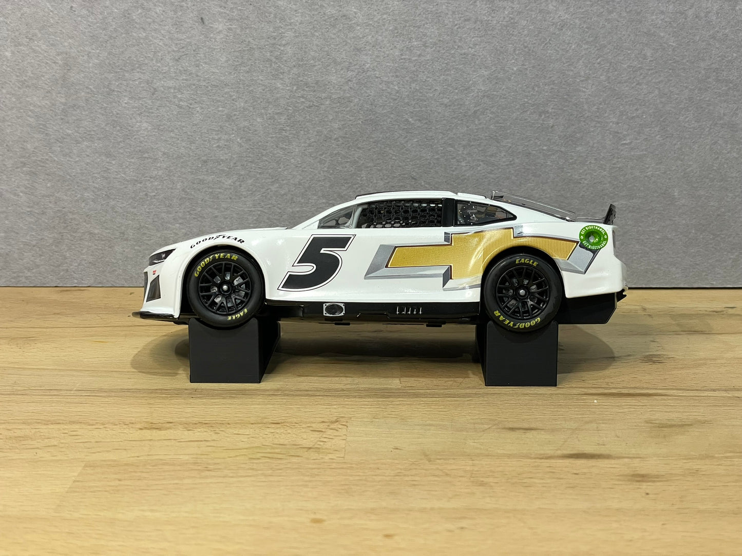 A 1:24 scale model of a NASCAR car displayed on two black angled display mounts on a wooden surface against a grey background.