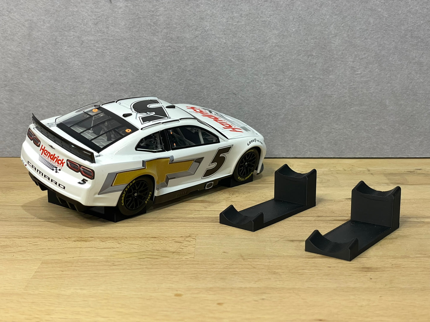 1:24 scale NASCAR diecast model with angled display mounts, featuring a white Hendrick Motorsports Chevrolet Camaro #5 on a wooden surface beside black support stands.